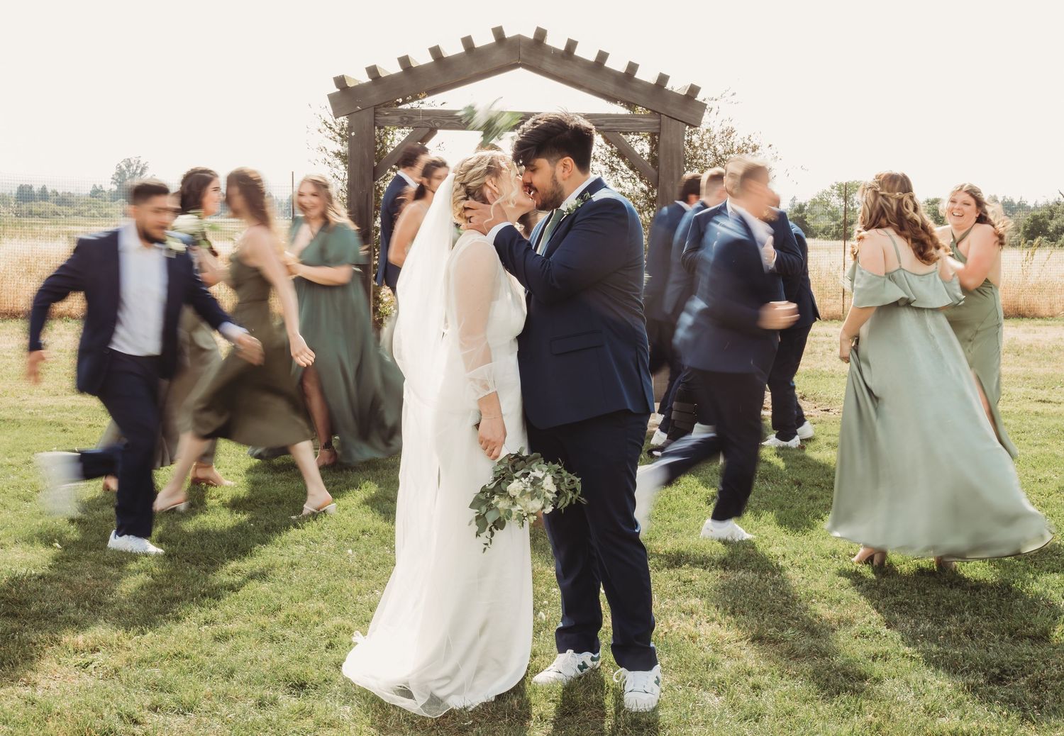 Wedding party dressed in suits and gowns walks through a grassy field during a sunny outdoor ceremony.