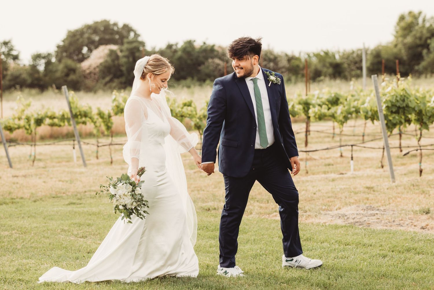 A couple walks hand in hand in a vineyard setting during a wedding celebration on a sunny afternoon.