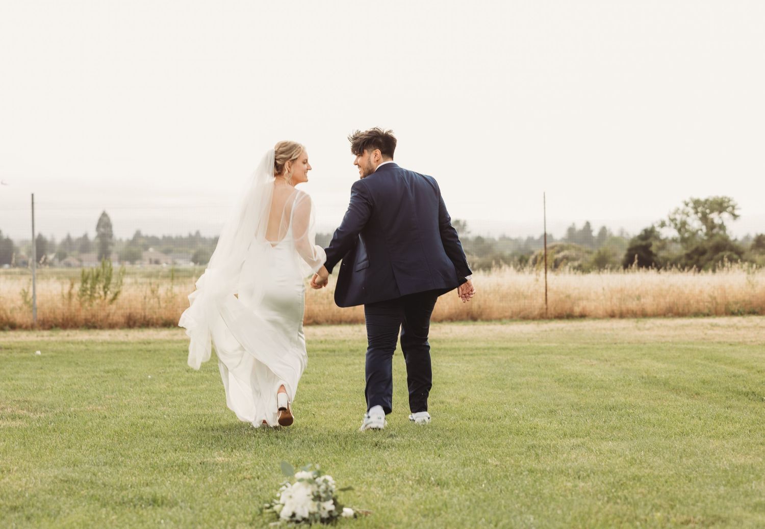 A couple runs together across a grassy field on their wedding day, with the bride's white dress flowing in motion.