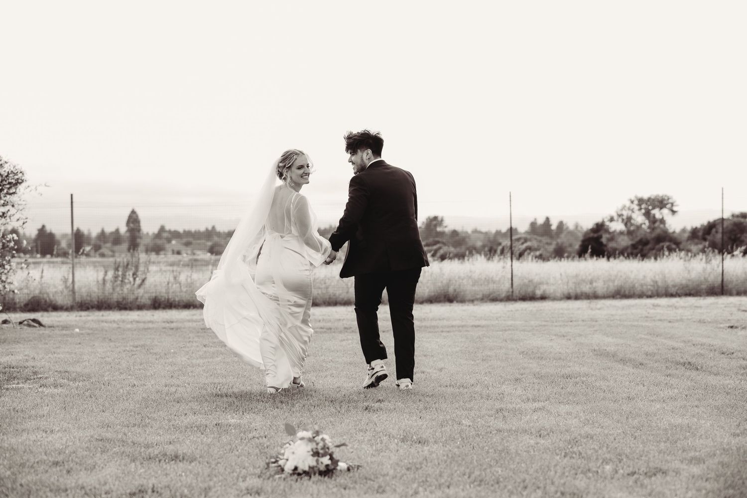 A couple in wedding attire walk together on a grassy field, with a small bouquet left behind on the ground.