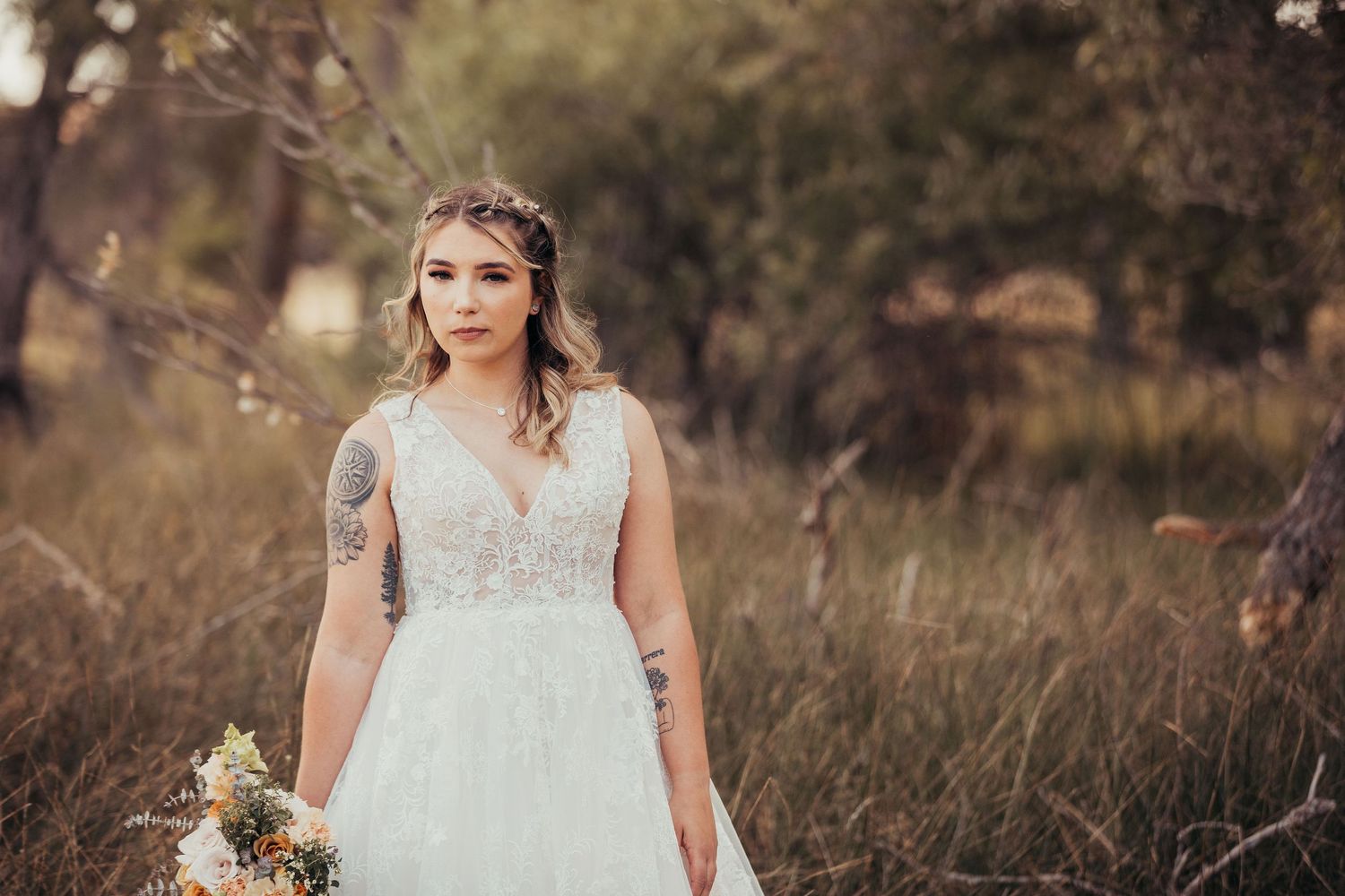 A bride in a lace V-neck wedding dress holds a bouquet while standing in a sunlit rustic field at dusk.