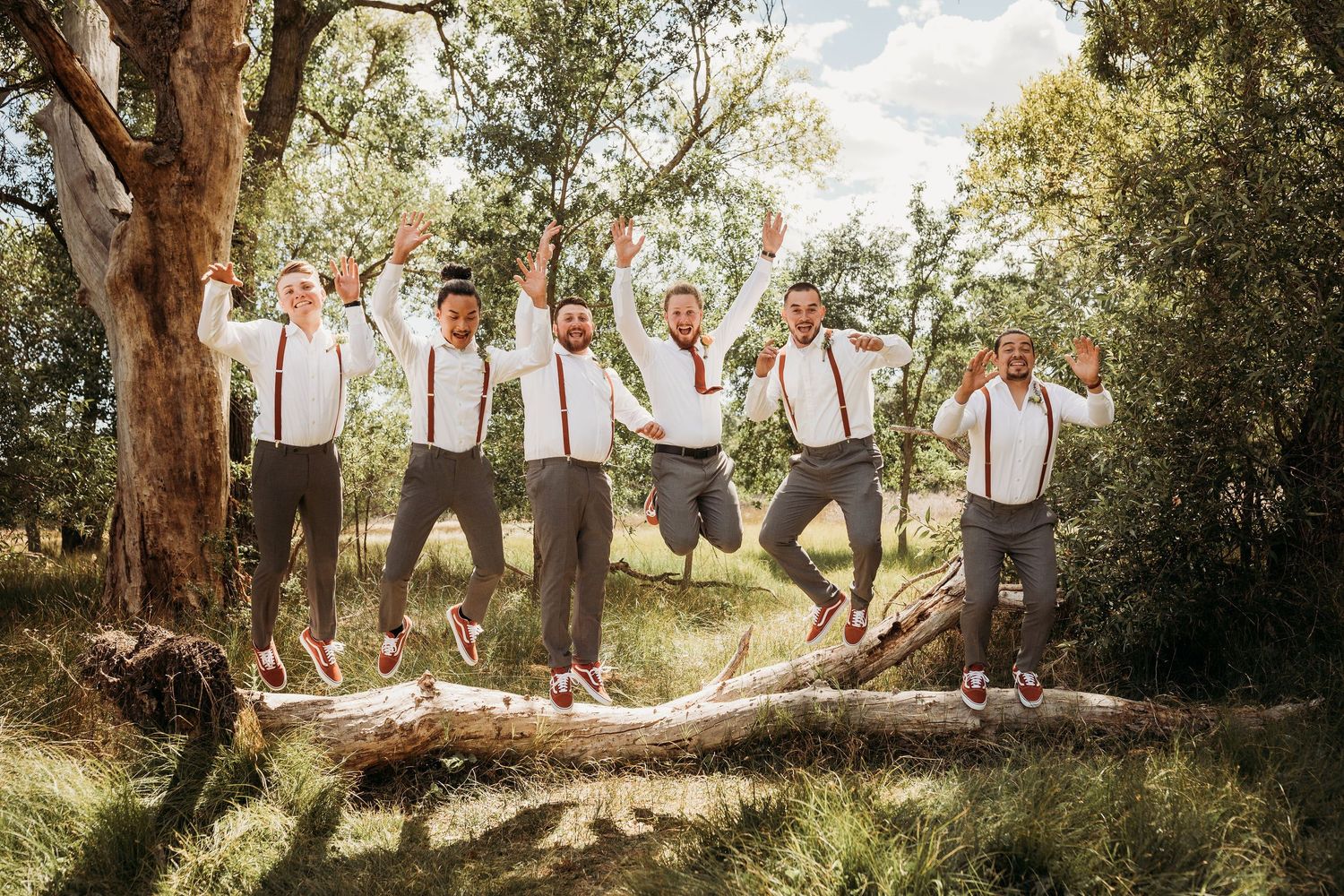 Groomsmen in matching outfits and suspenders pose playfully while jumping in unison outdoors in a wooded setting.