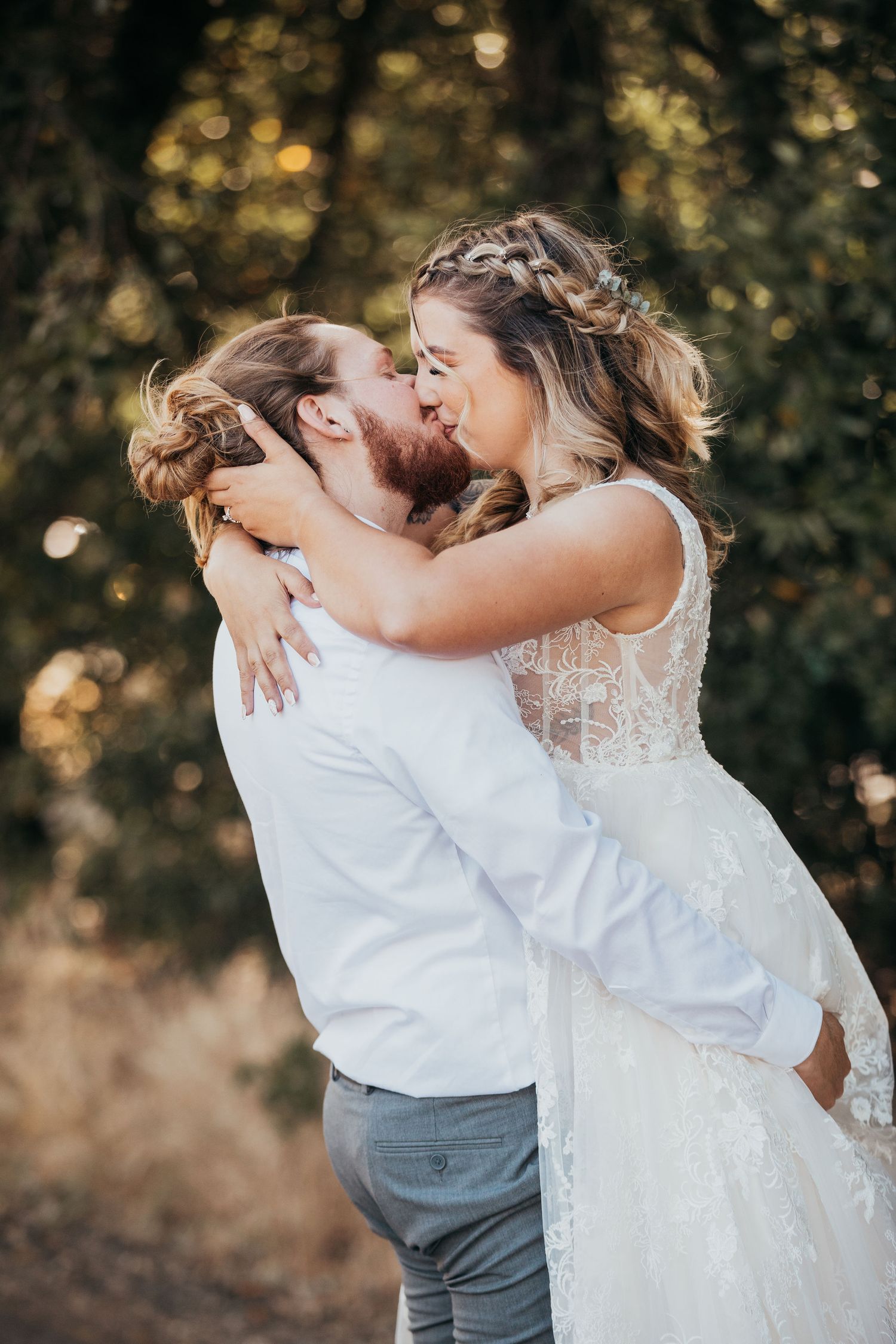 Newlywed couple shares a passionate kiss outdoors with sunlight filtering through trees in romantic wedding photos.
