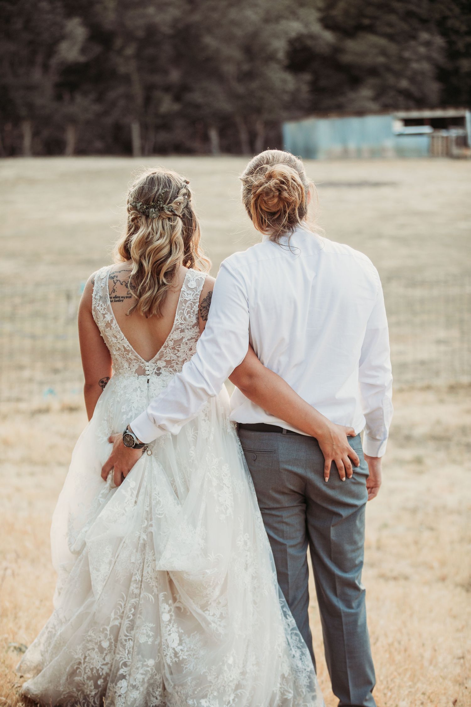 A couple walks together in a natural field setting with a bride wearing a lace dress and her partner in gray attire.