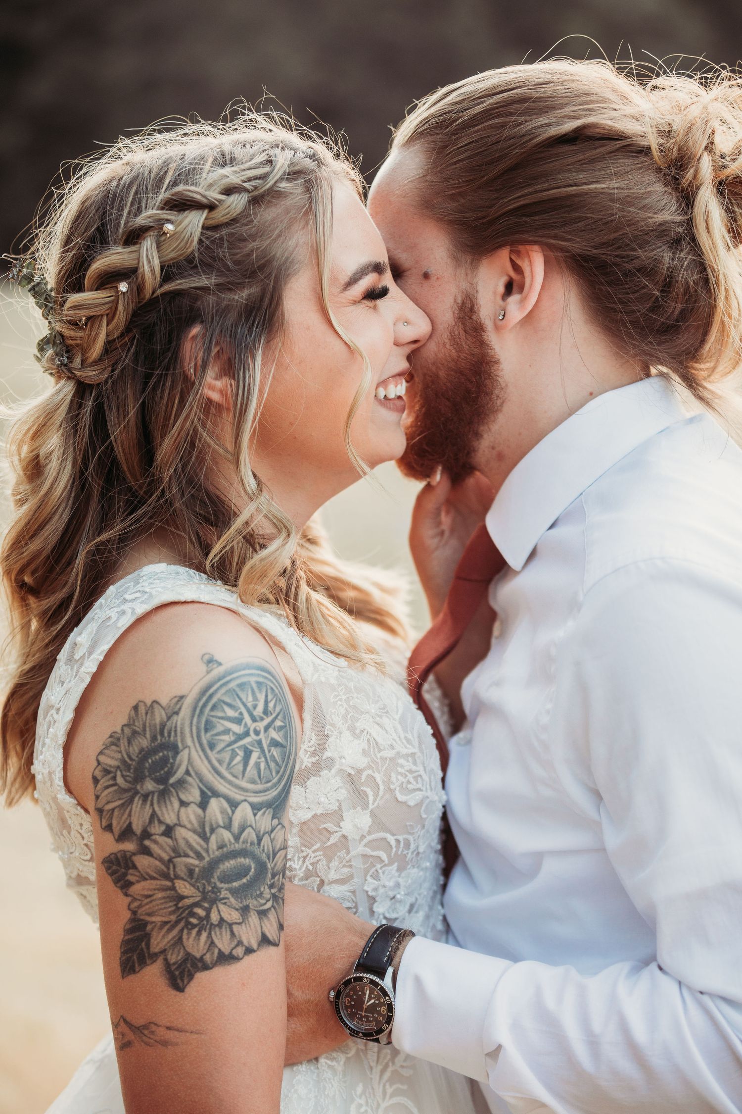 A romantic wedding couple share an intimate moment, showcasing the bride's floral arm tattoo and braided hairstyle.