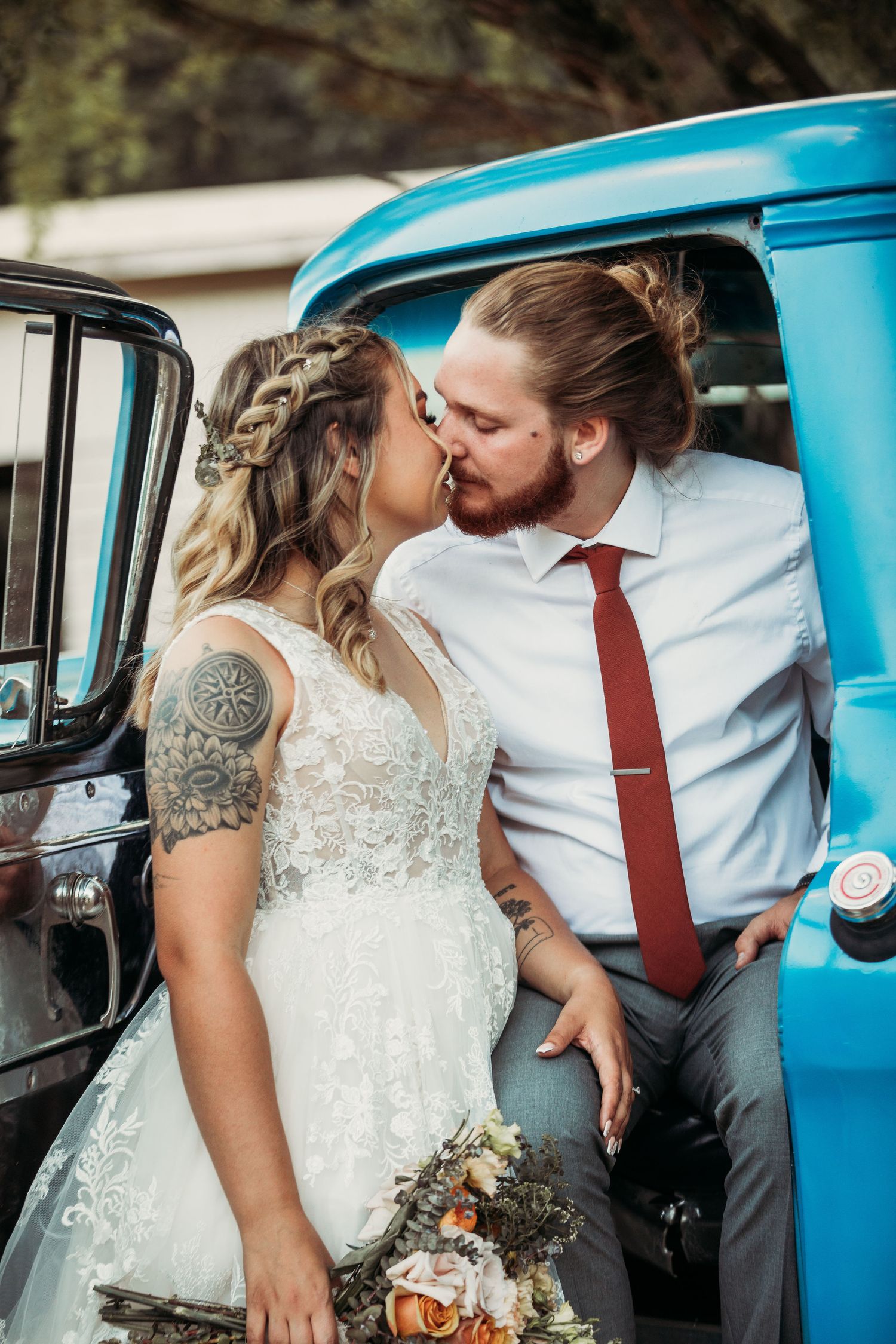 A newlywed couple poses against a vintage blue truck, with the bride wearing a lace dress and holding a bouquet.