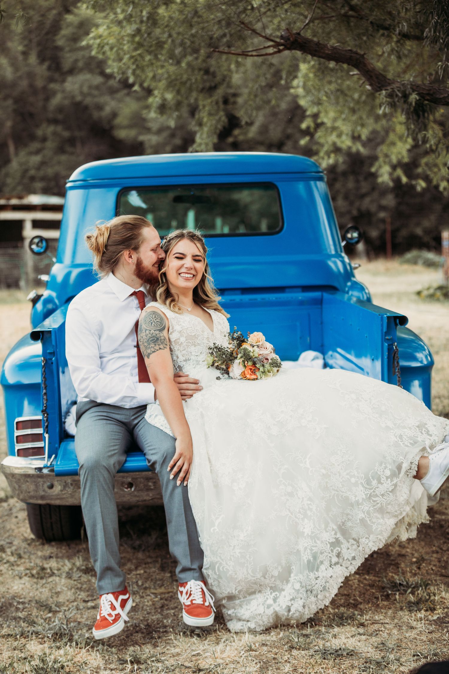 A joyful couple sits on the bed of a bright blue vintage pickup truck during their rustic wedding photoshoot.
