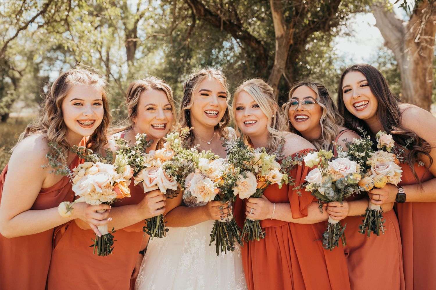 Wedding party in matching rust-colored bridesmaid dresses pose together holding neutral-toned floral bouquets outdoors.