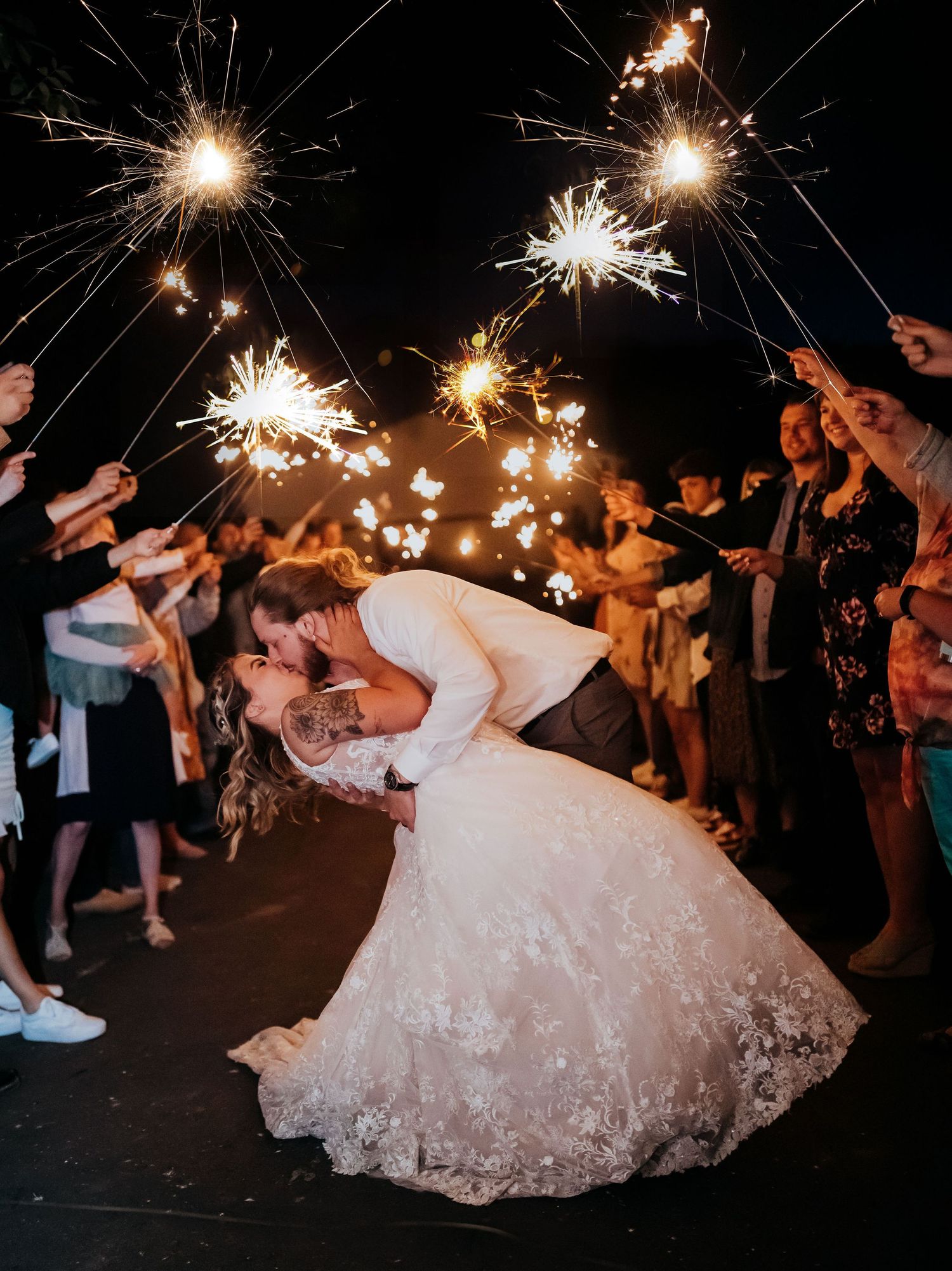 Couple shares romantic dip kiss under glowing sparklers held by wedding guests during nighttime celebration.