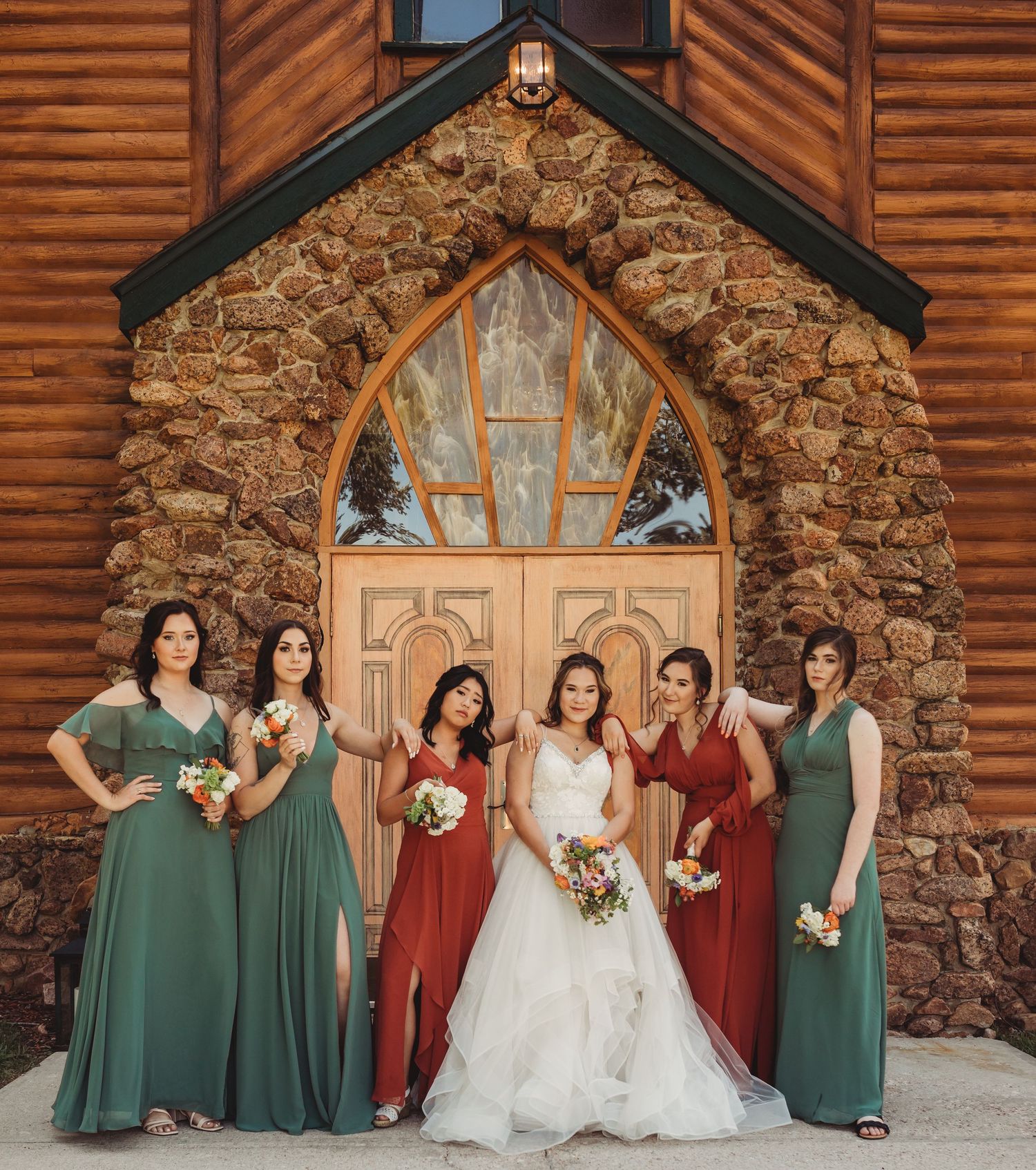 Bridal party in alternating dark green and burgundy dresses pose in front of a rustic stone and wood lodge.