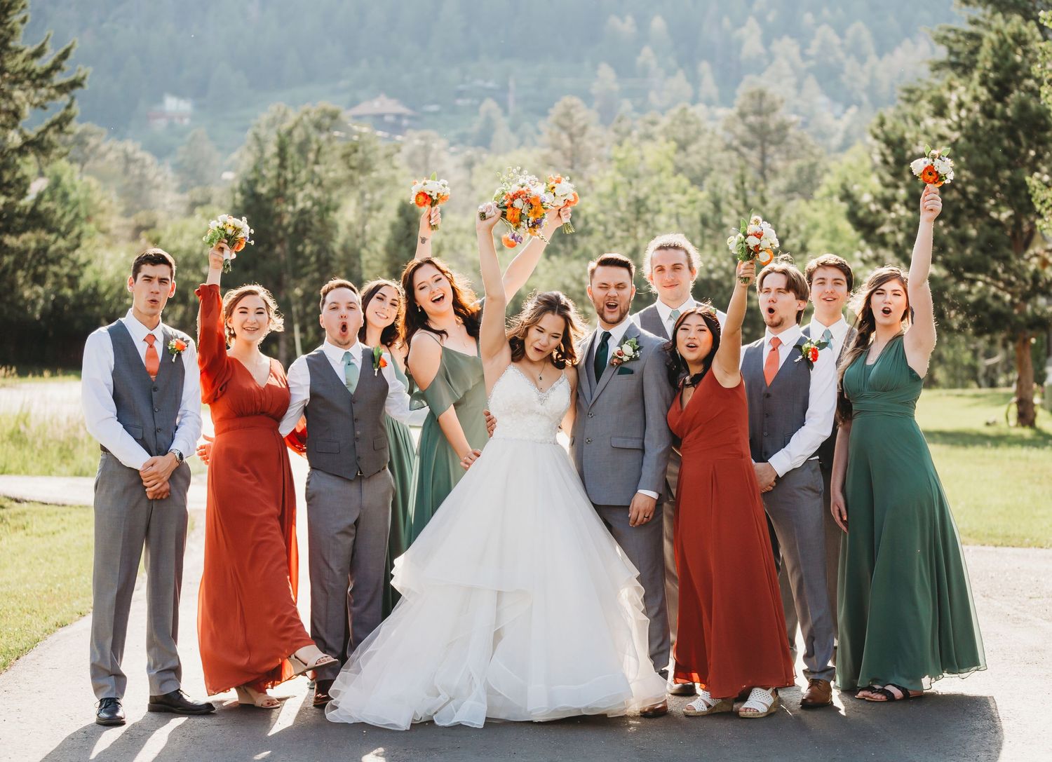 Wedding party wearing colorful red and green dresses and gray suits poses together outdoors in summer landscape.