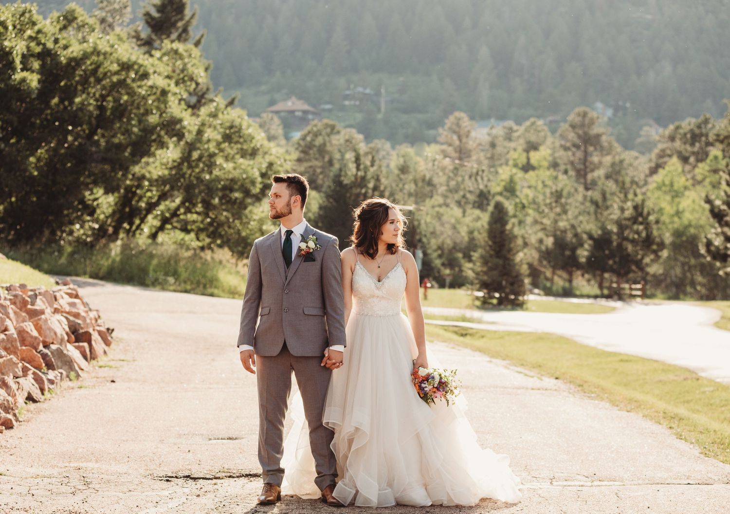 A beautiful wedding couple walks down a sunlit dirt path surrounded by lush greenery and natural scenery.