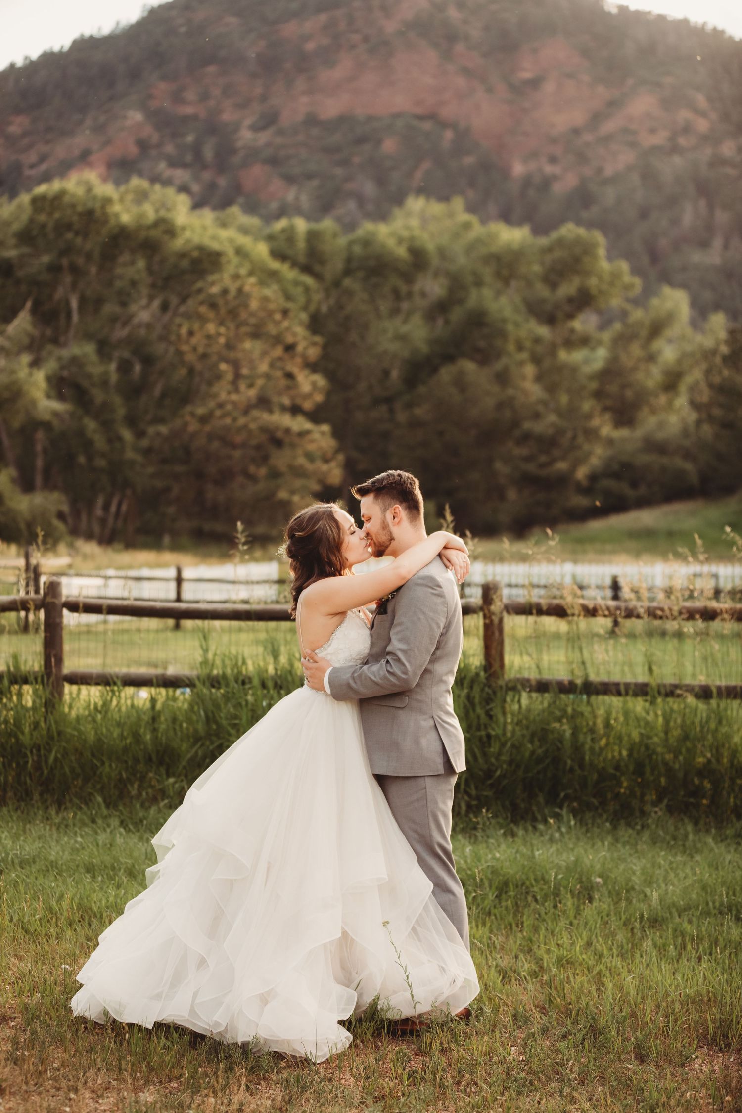 A romantic wedding couple embraces in a rustic field setting with mountains and trees in the background at sunset.