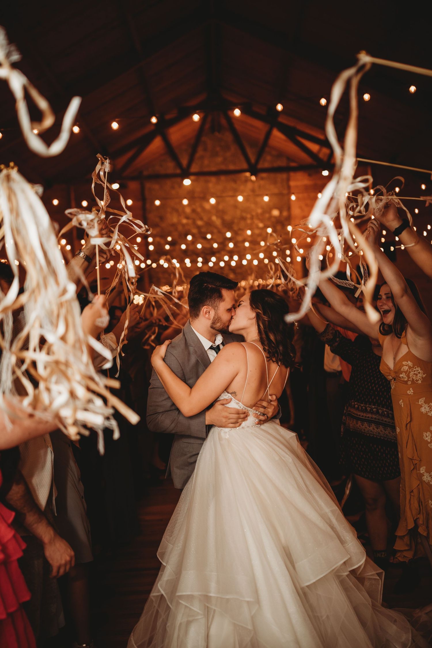 Newlyweds share a romantic dance under twinkling string lights at a rustic barn reception while guests wave streamers.
