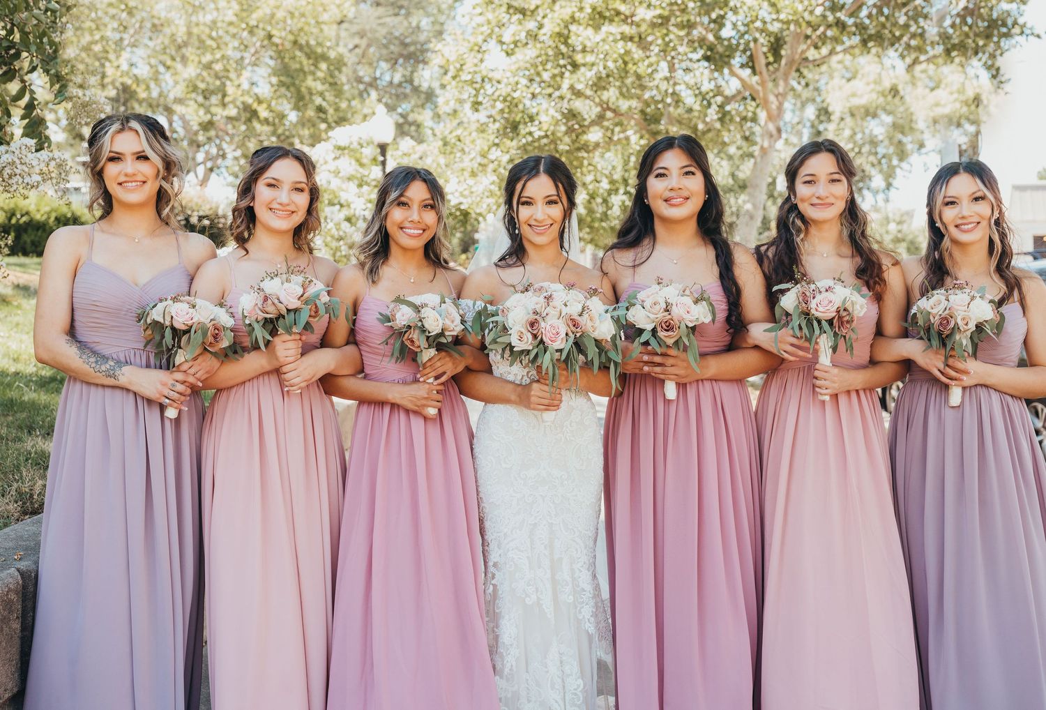 Bridesmaids in lavender and mauve dresses stand together holding white floral bouquets during an outdoor wedding.