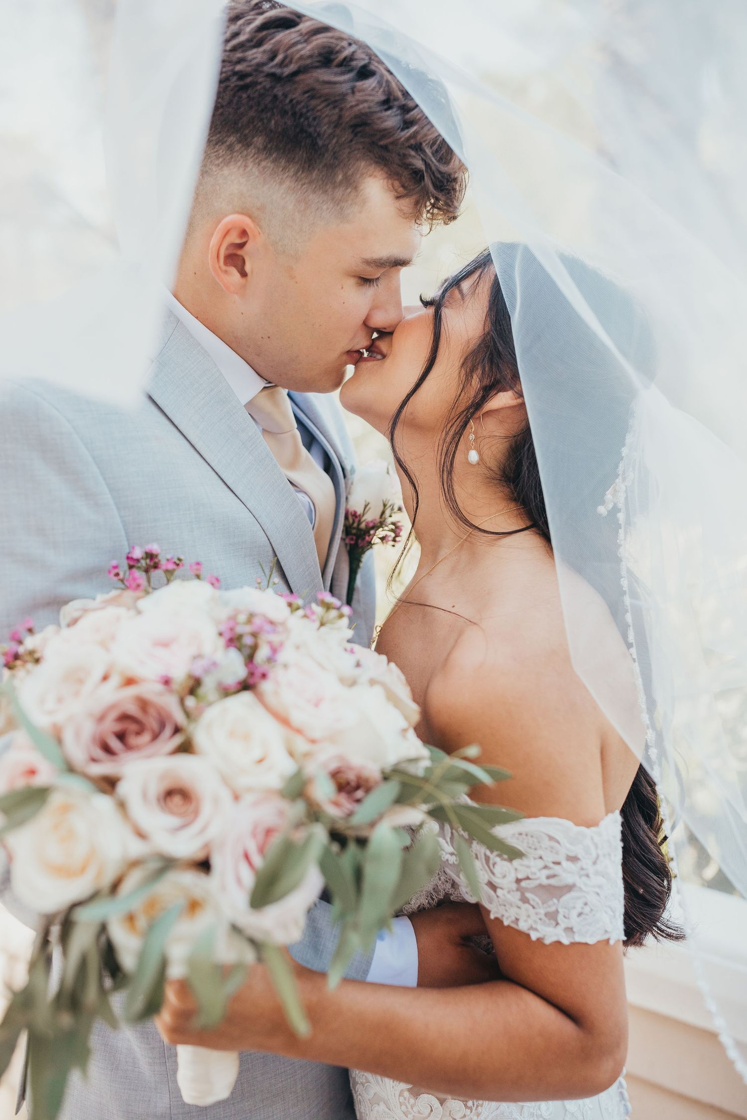Romantic wedding photo featuring a tender kiss under a delicate bridal veil with blush roses and eucalyptus bouquet.
