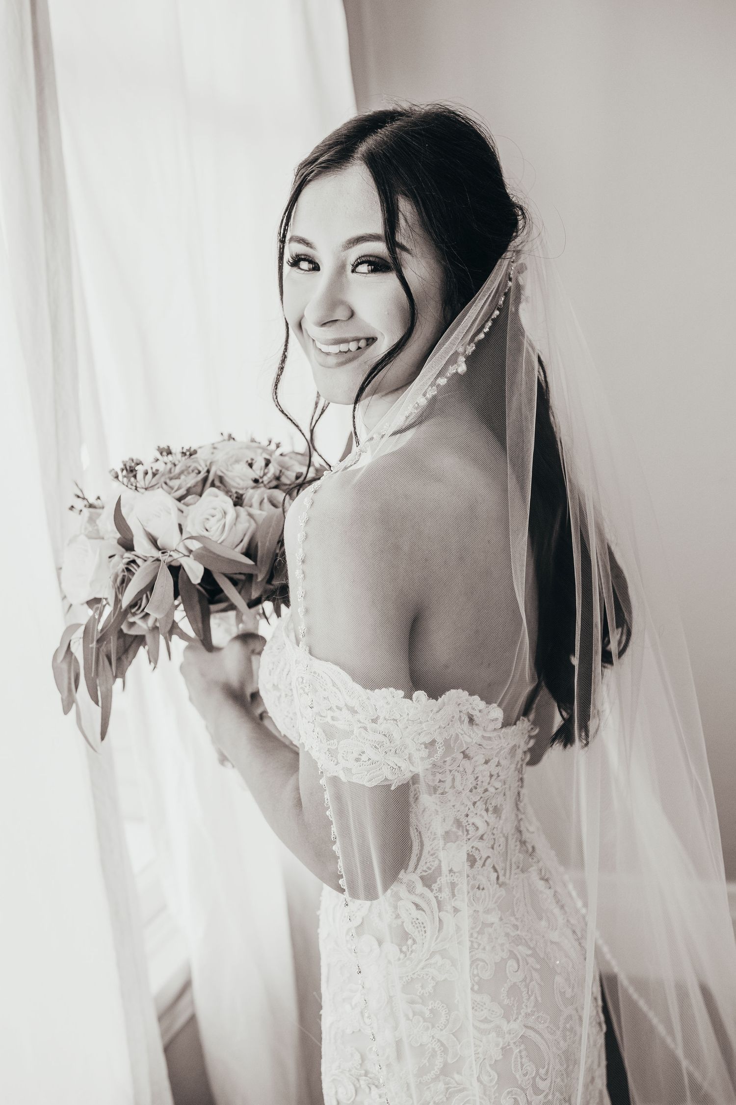 Beautiful bride in off-shoulder lace wedding dress and veil holding floral bouquet poses with natural smile during bridal shoot.