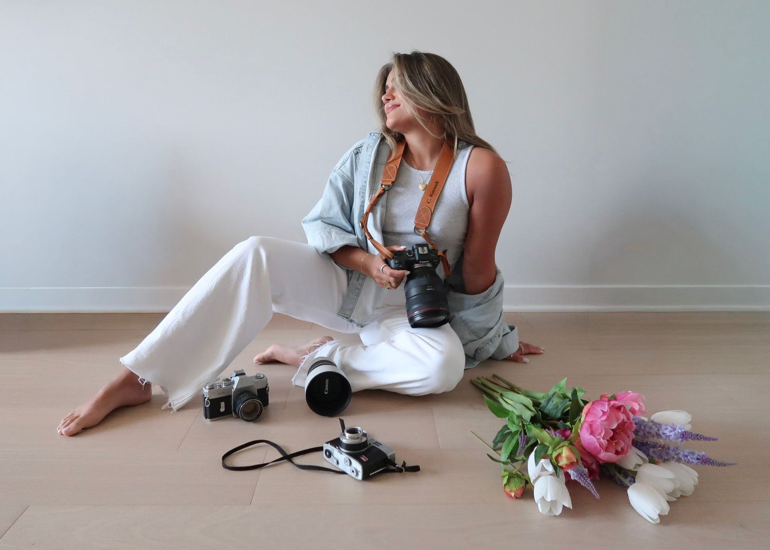 A photographer in white loungewear sits on floor reviewing camera images with photography equipment and flowers nearby.