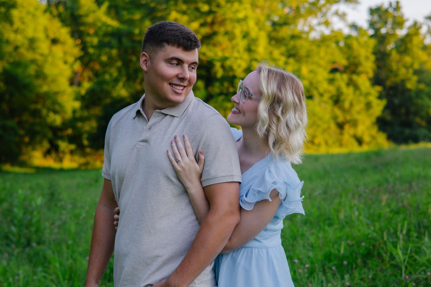 Two people share a tender moment while standing in a sunlit field with golden foliage behind them.