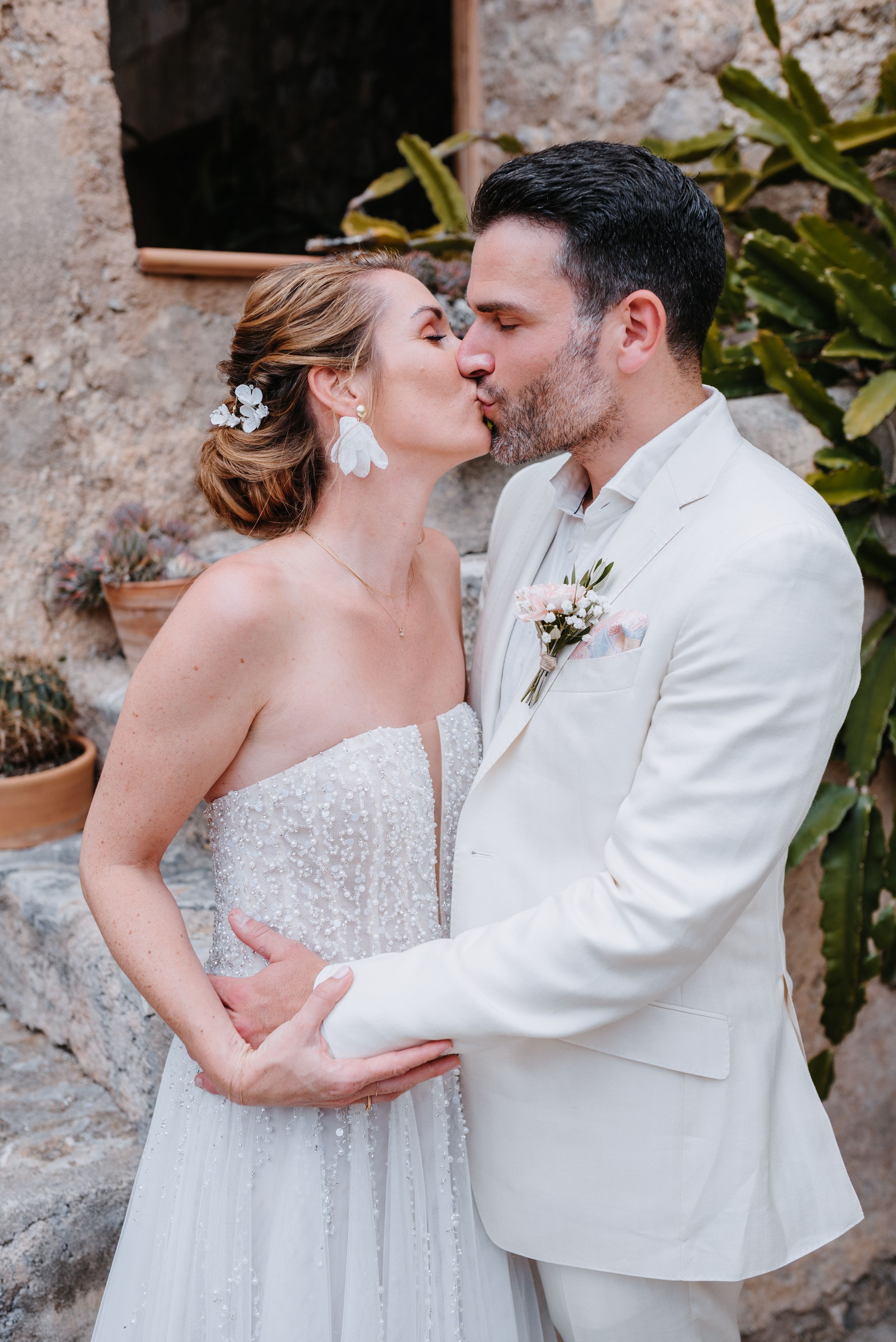Ein romantisches Hochzeitspaar, das einen zärtlichen Moment und einen Kuss im Freien vor einer mallorquinischen Steinmauer teilt.