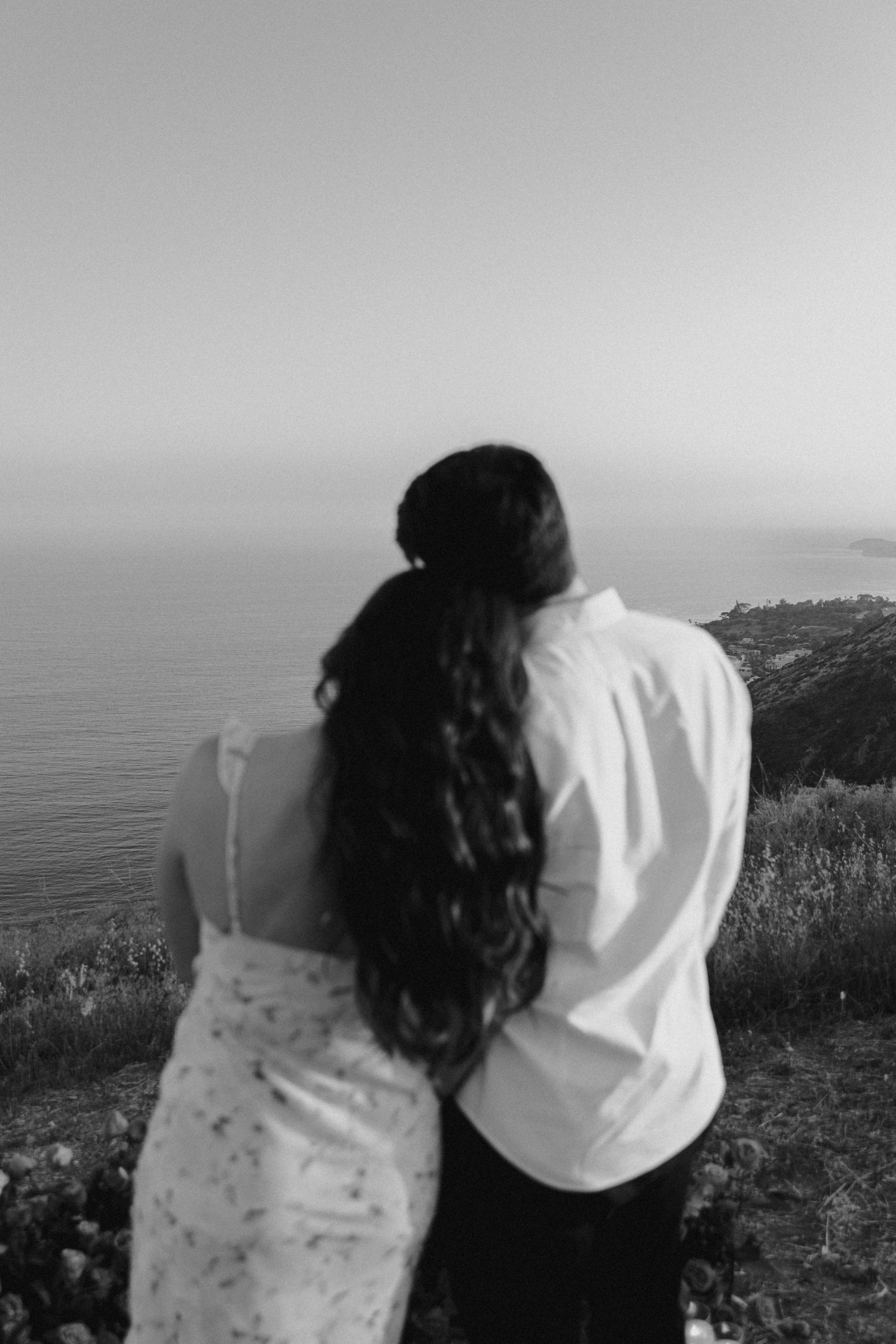 Black and white photo of a couple embracing while overlooking an ocean vista from a cliff during a scenic moment.