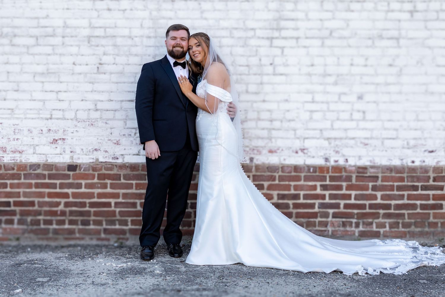 A bride and groom pose together against a rustic white brick wall with contrasting red brick at the bottom.