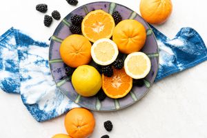 Oranges, blackberries and lemons arranged on a purple plate on top of a tie dye blue and white napkin.