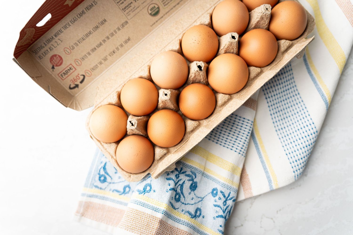 A dozen brown eggs arranged in an open cardboard carton resting on a blue and yellow striped kitchen towel, ready for cooking or baking.