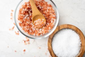 Coarse pink Himalayan salt crystals in a clear glass bowl with a wooden spoon, next to a small wooden bowl of fine white sea salt.