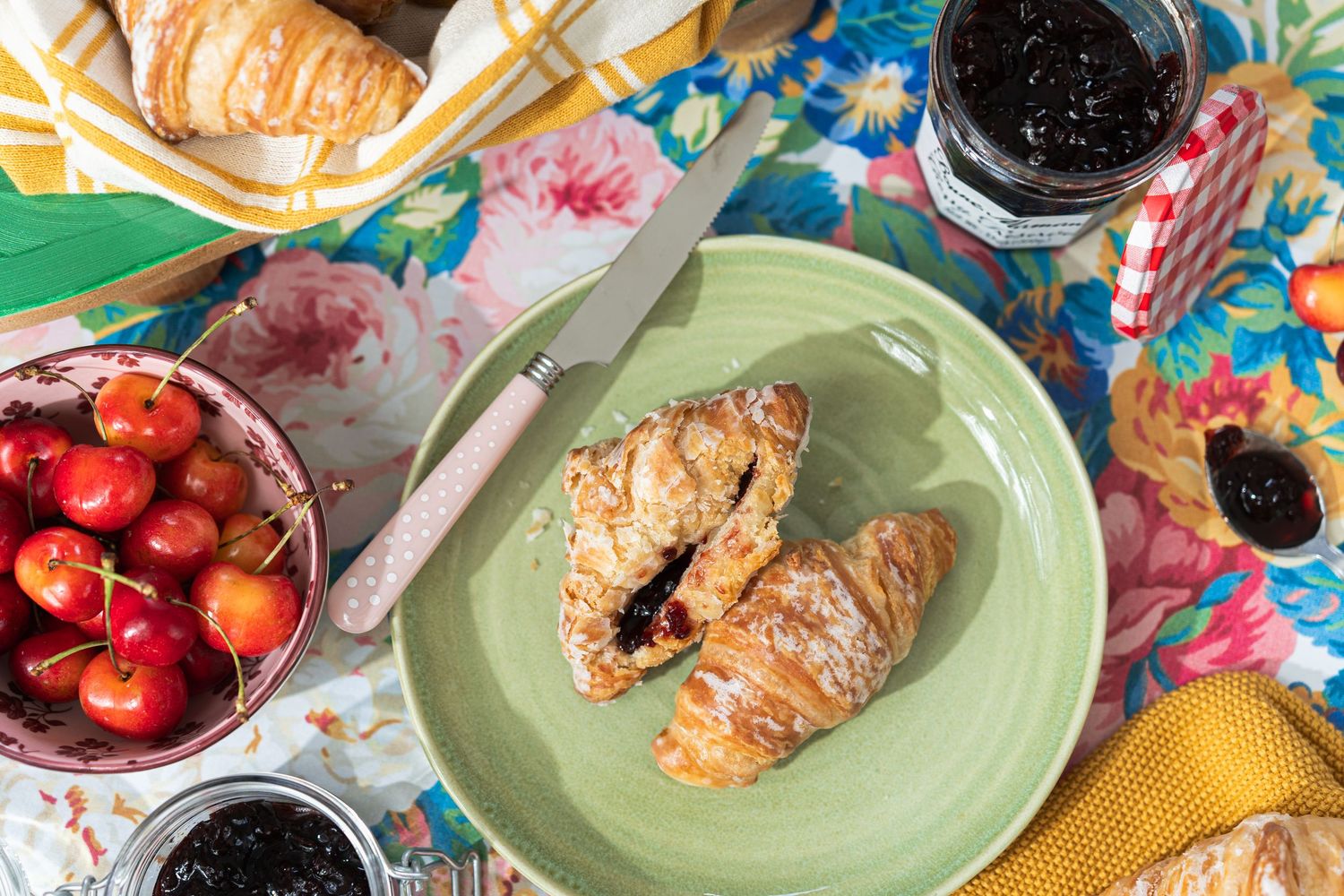 Flat lay of croissant with jam sits on a green plate with a coquette style knife. Jars of jam and fresh cherries sit on a floral backdrop.