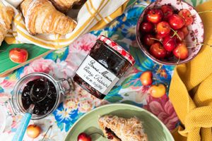 Flat lay of croissant with jam sits on a green plate with a coquette style knife. Jars of jam and fresh cherries sit on a floral backdrop.