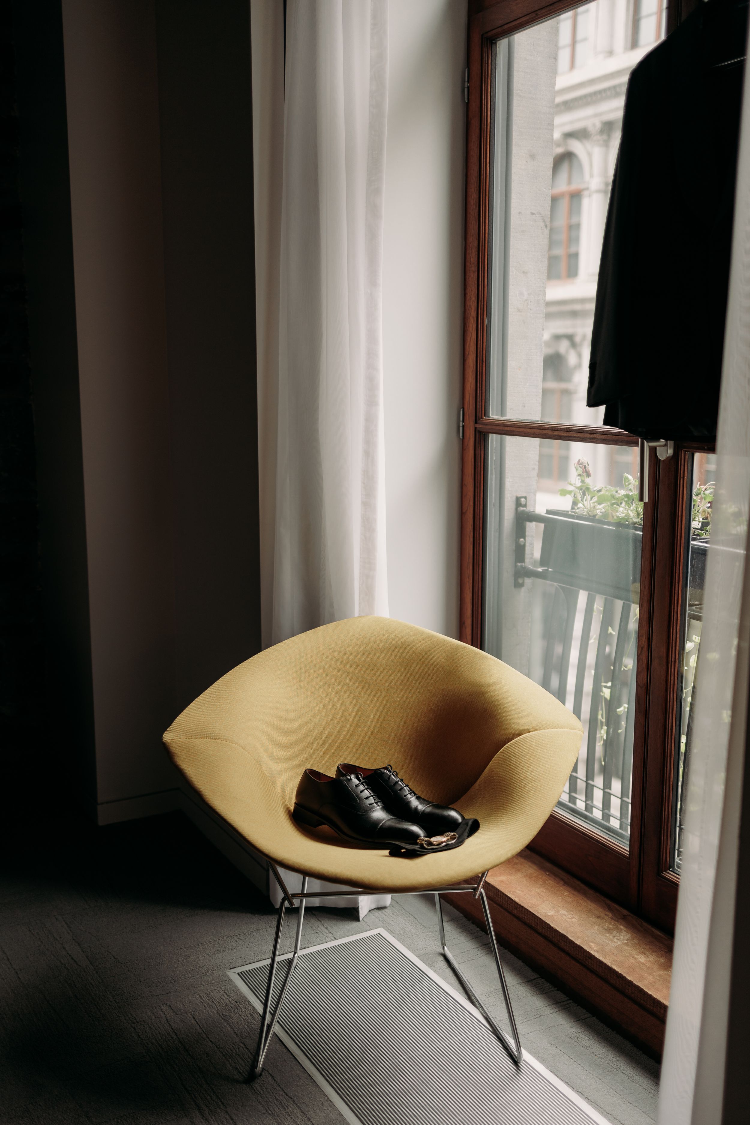 A pair of black formal shoes placed on a beige modern chair by a window with white curtains.