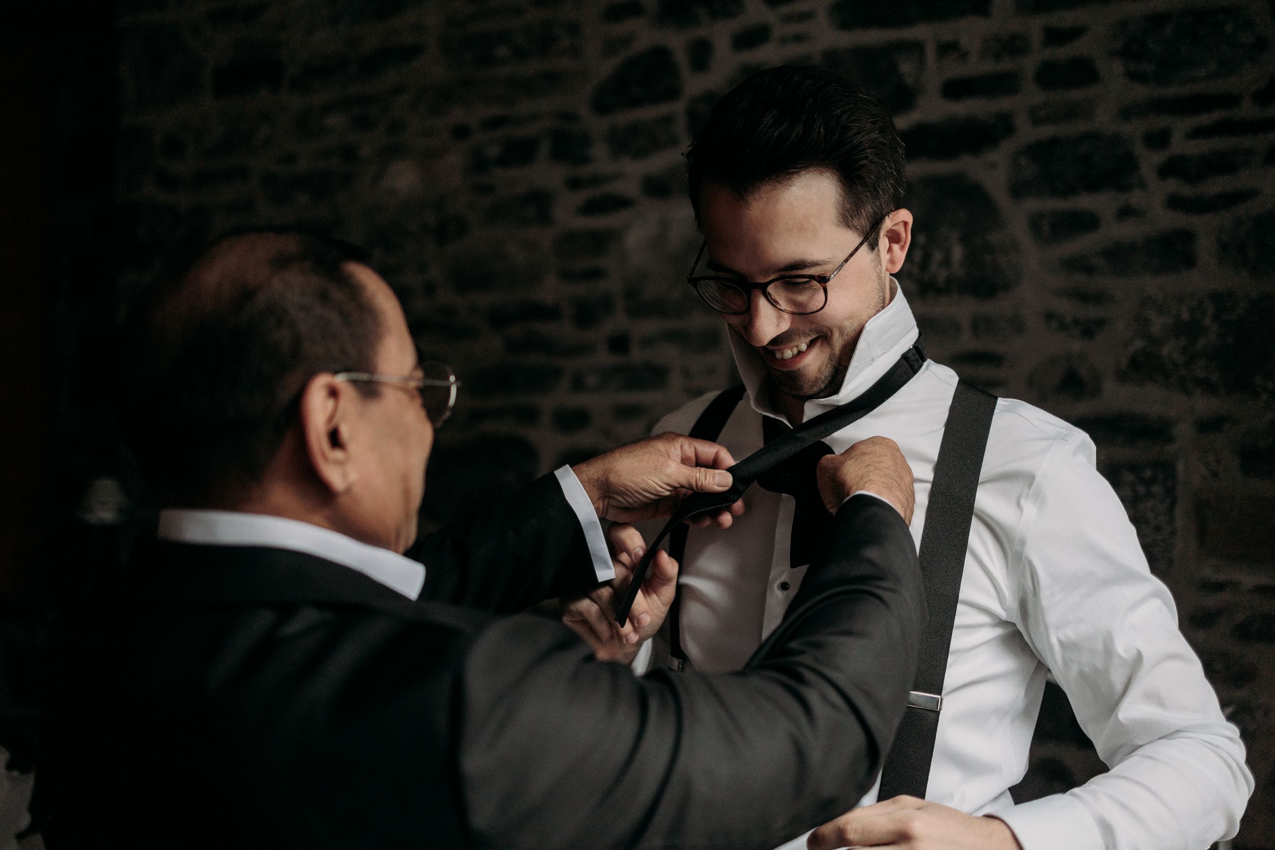 Two people in formal attire help adjust a bow tie against a dark stone wall background.