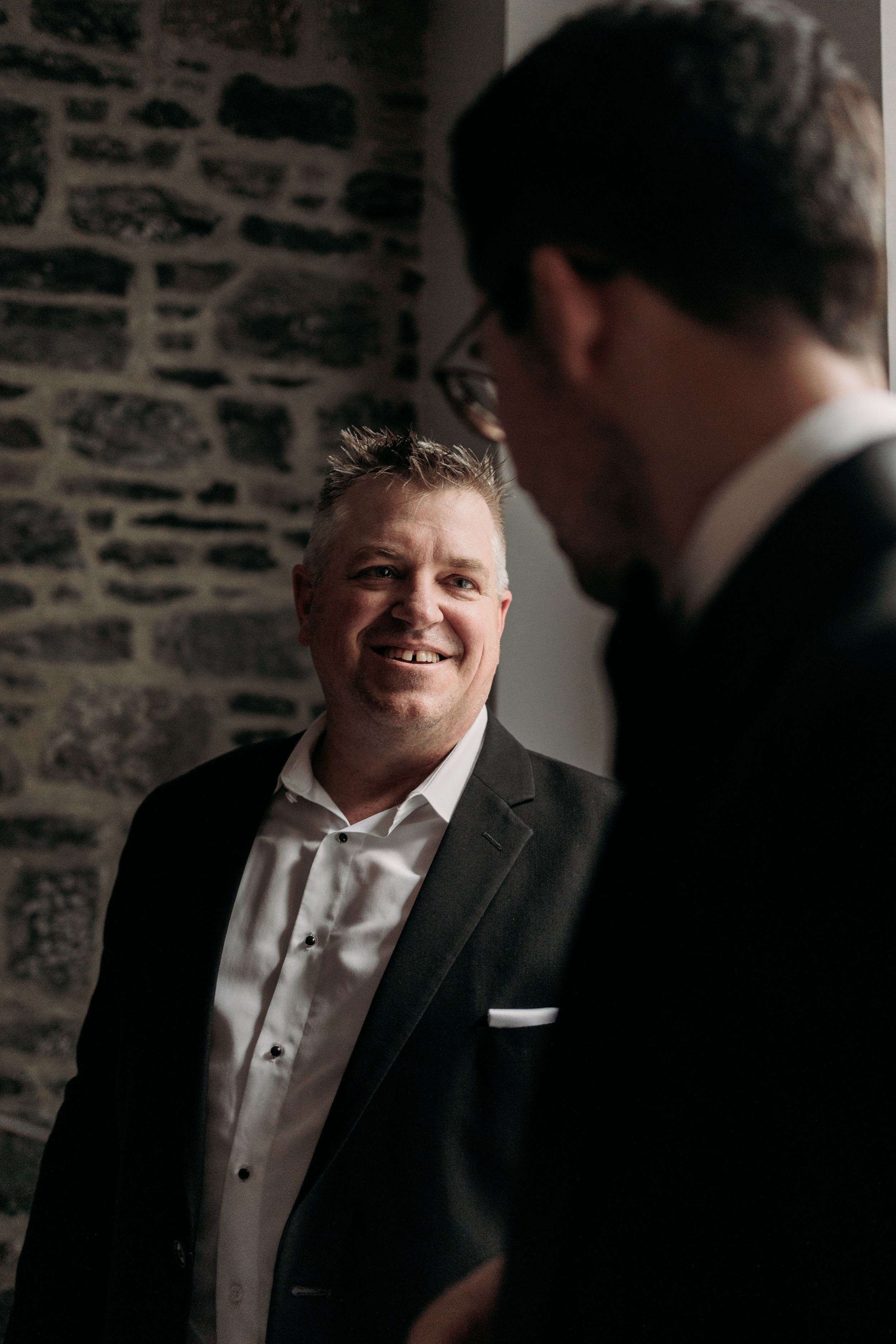Groom in black tuxedo laughs while standing against rustic stone wall during wedding.