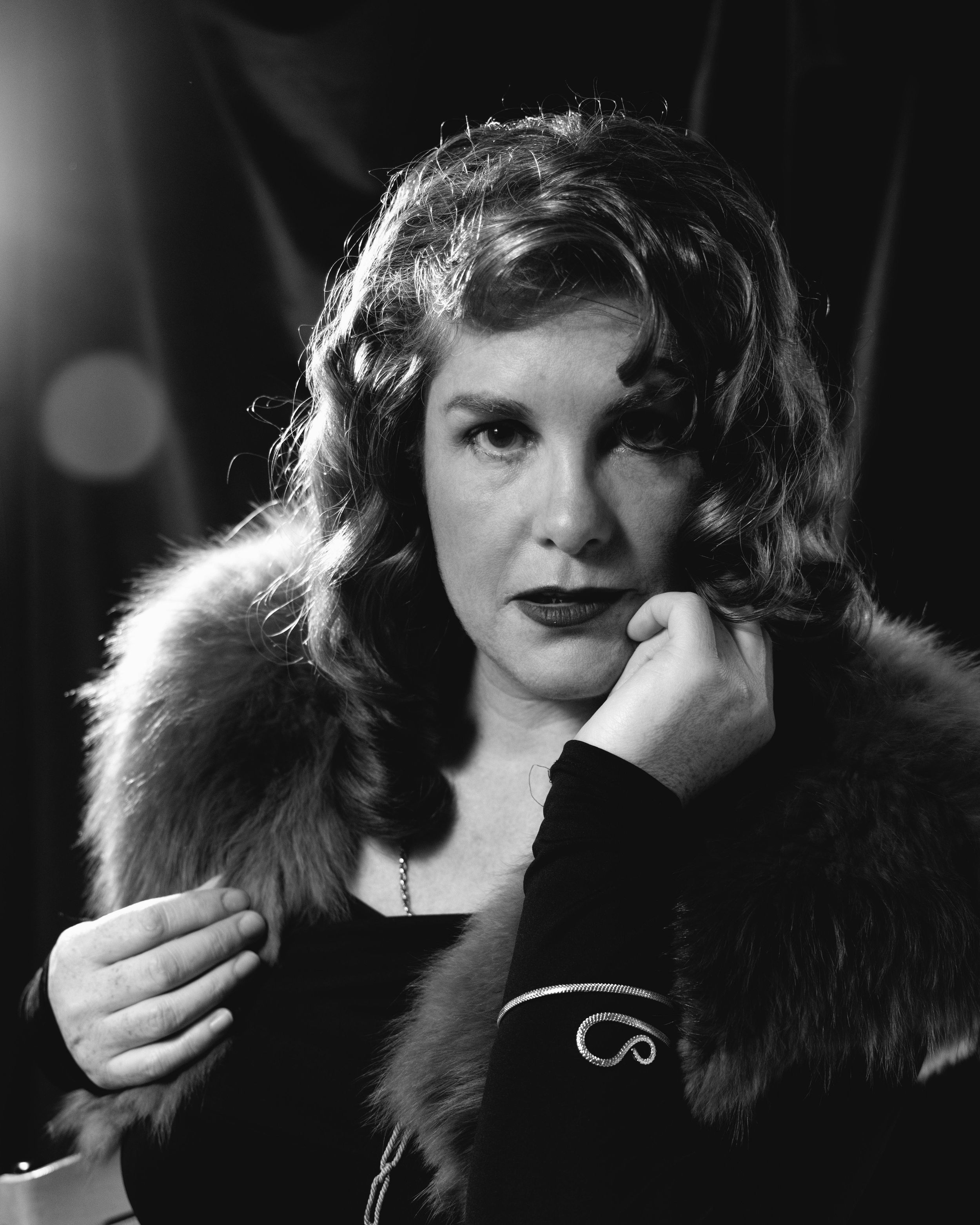 Black and white portrait of a woman in a fur coat, resting her chin on her hand with dramatic lighting.