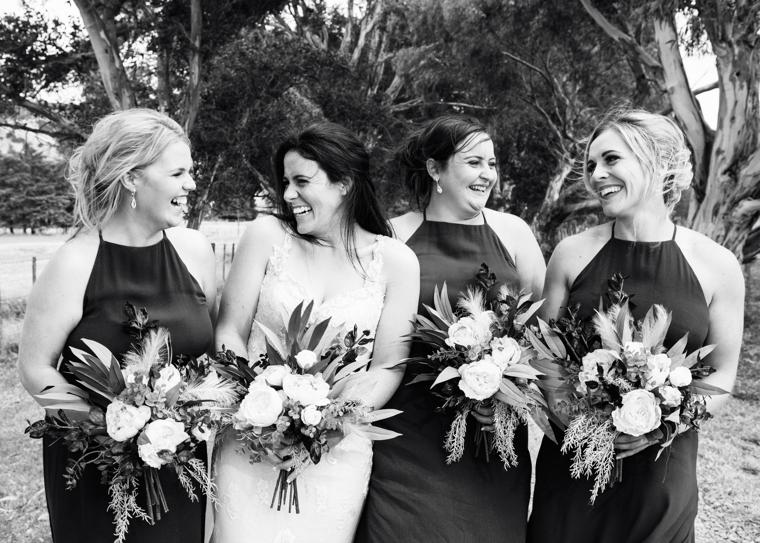 A joyful bridal party in black dresses shares a candid moment while holding elegant floral bouquets at an outdoor venue.