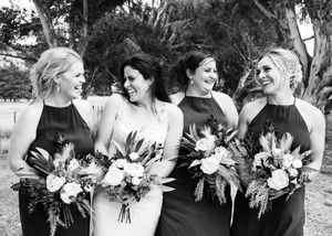 A joyful bridal party in black dresses shares a candid moment while holding elegant floral bouquets at an outdoor venue.