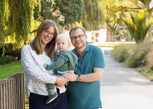 A family of three smiles together on a pathway with drooping willow trees and lush greenery in the background.