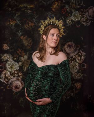 Dark moody maternity portrait with an ornate gold crown against a floral backdrop, wearing emerald velvet gown.