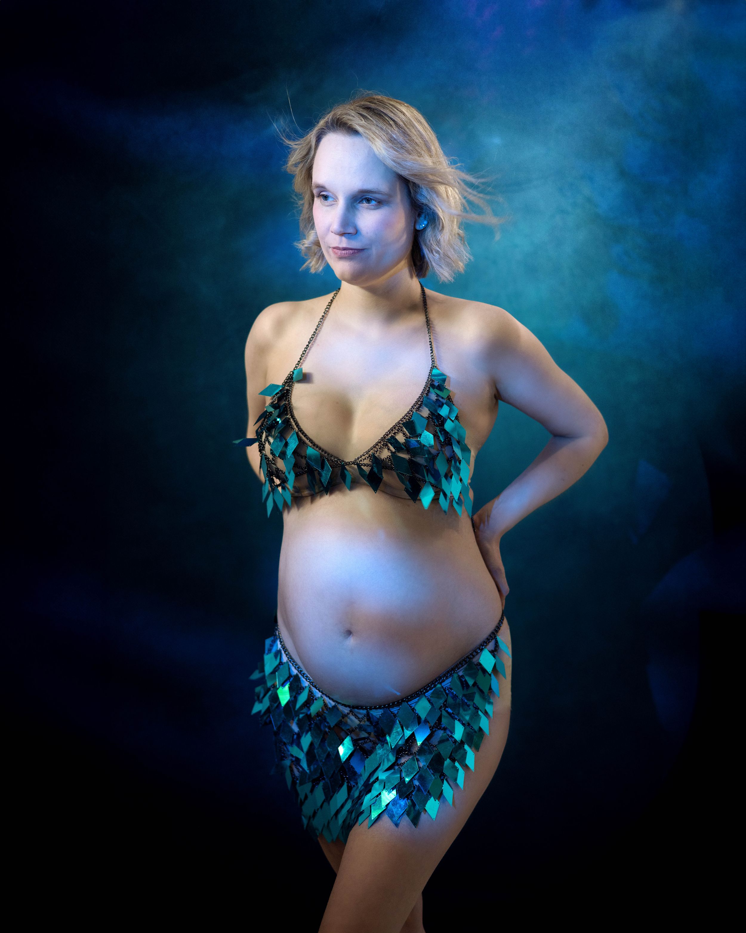 A maternity photography series with moody blue lighting showcases a woman posing in a teal butterfly-adorned outfit.