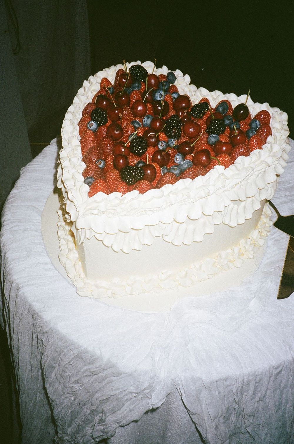 Heart-shaped white frosted cake decorated with fresh strawberries and blueberries on a white tablecloth.