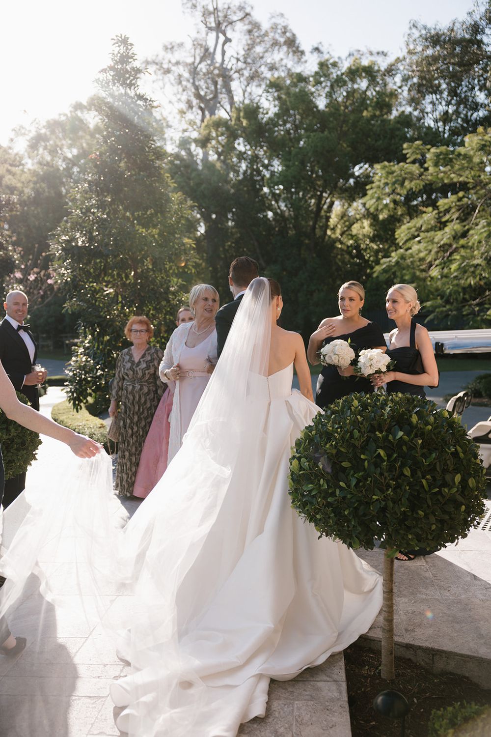 Sequence of bridal party helping with the train of a flowing white wedding dress in an outdoor garden setting.