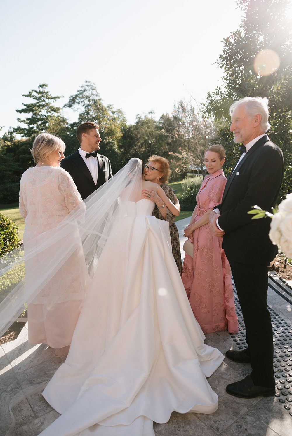 Wedding party walks together along a sunlit pathway with elegant flowing white gown trailing behind them.