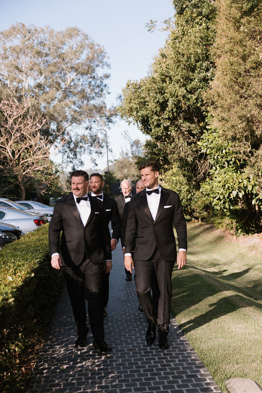 Groomsmen walking and laughing together along outdoor garden path in black tuxedos before wedding ceremony.