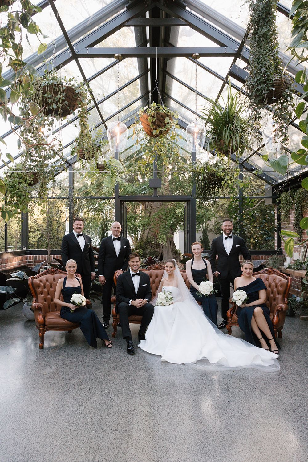 Wedding party relaxes in an elegant greenhouse conservatory with glass ceiling and lush greenery.