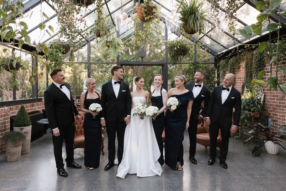 Wedding party members laughing and posing together under a glass greenhouse ceiling for formal photos.