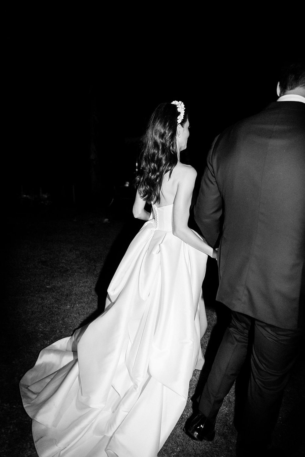 A dramatic black and white photo captures a bride in flowing white gown walking with her partner at night.