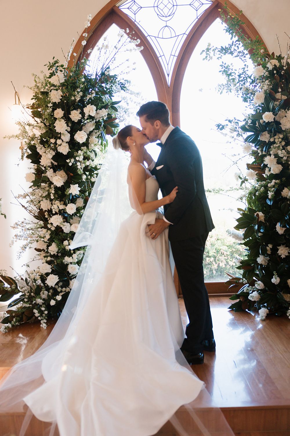 A romantic wedding couple shares their first kiss in a chapel with elegant floral arrangements framing the scene.