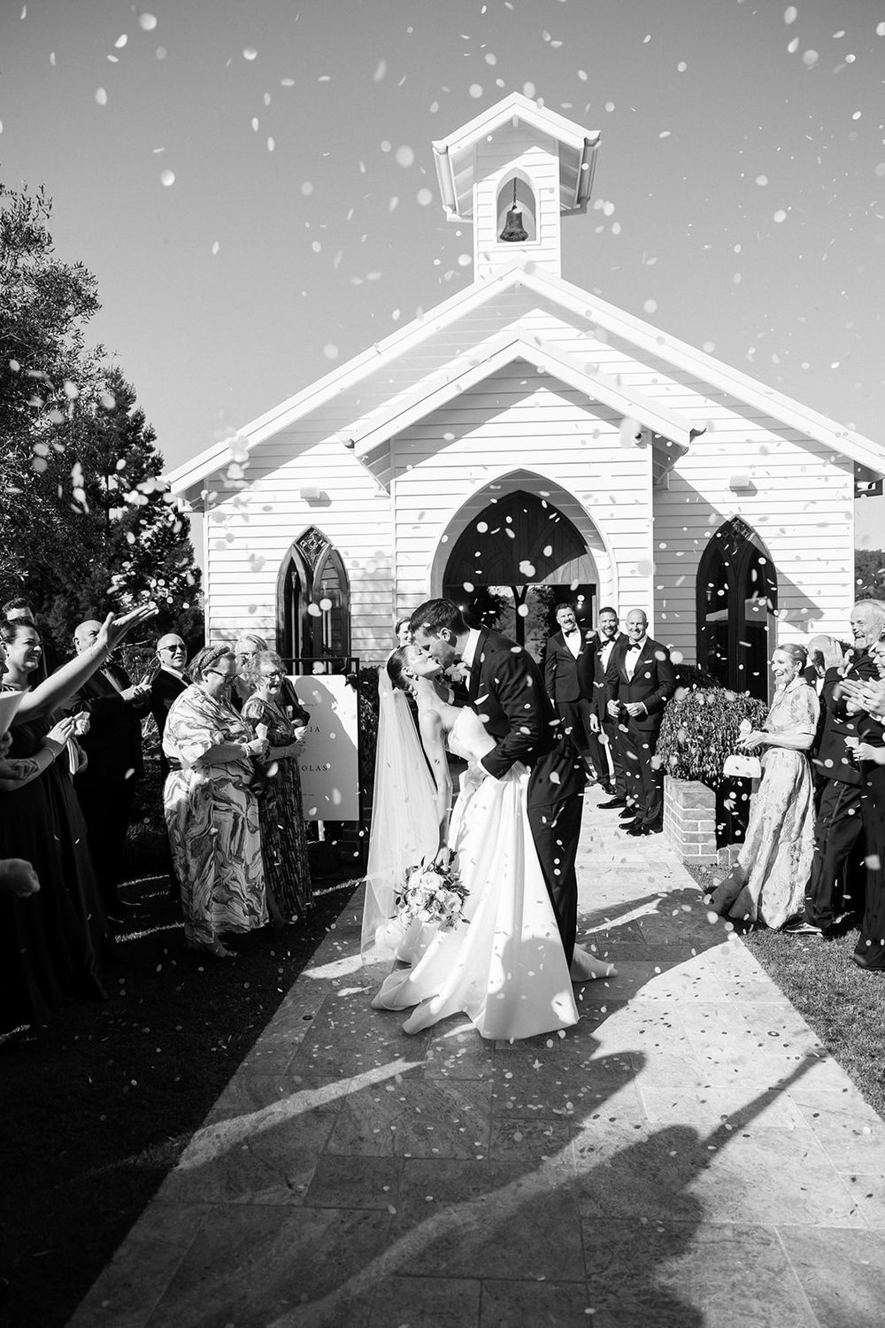 A joyous celebration outside a charming white chapel as wedding guests throw confetti in this black and white photograph.