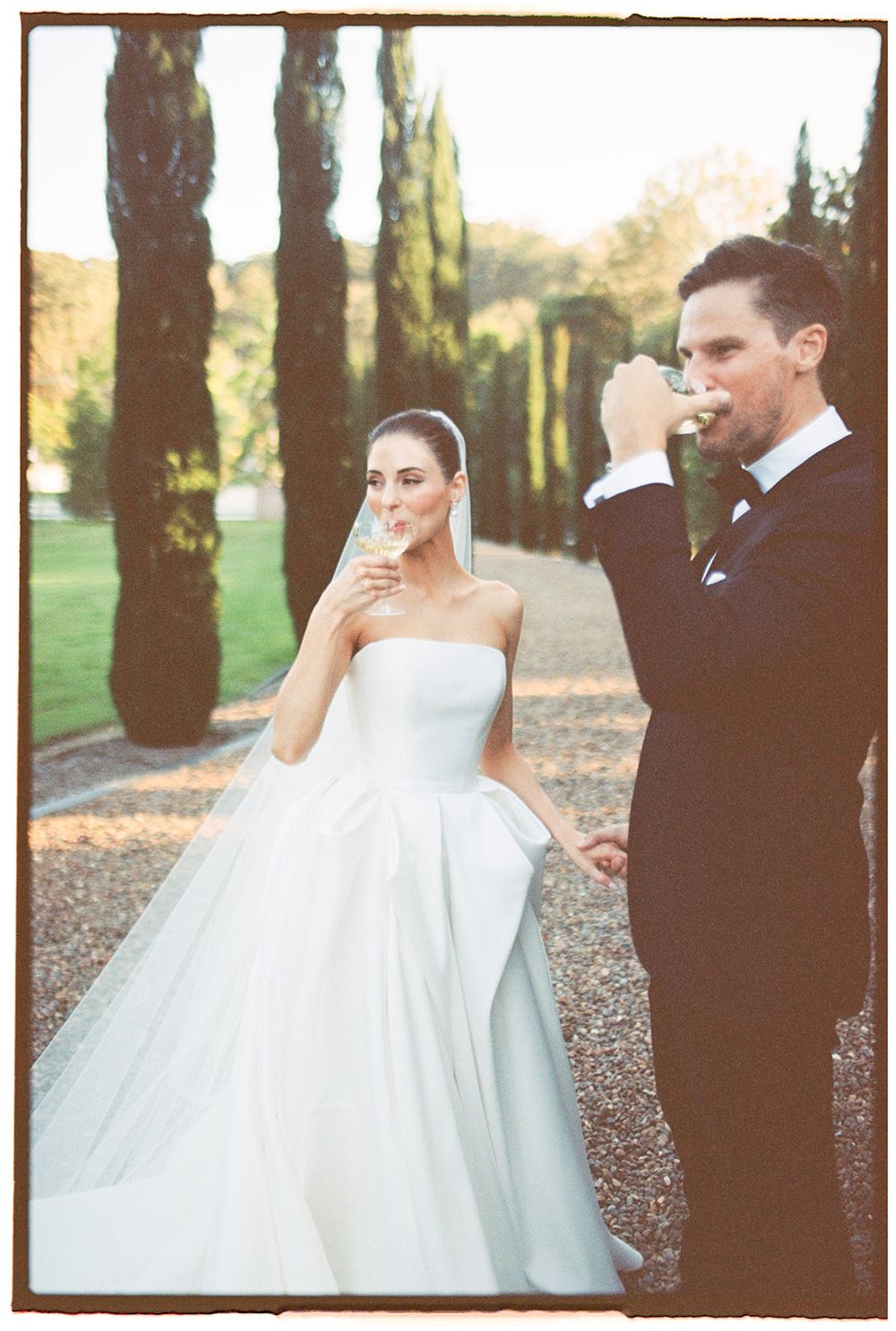 Romantic couple in wedding attire walks through a wooded path, captured in a dreamy vintage film style.